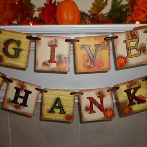 May include: A Thanksgiving banner with the words "Give Thanks" in brown letters on a beige background with fall-themed accents. The banner is strung with brown twine and has small pumpkins and leaves.