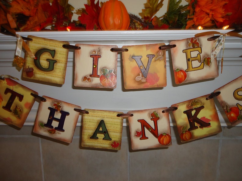 May include: A Thanksgiving banner with the words "Give Thanks" in brown letters on a beige background with fall-themed accents. The banner is strung with brown twine and has small pumpkins and leaves.