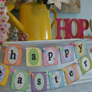 May include: A colorful banner with the words "Happy Easter" in a white oval on each of the individual banners. The banner is strung with white ribbon and colorful tulle. The banner is hanging on a white shelf with a yellow watering can, a pink "HOP" sign, and a green and yellow flower.