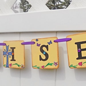 May include: A yellow and white banner with the words "He is Risen" in black letters. The banner is decorated with purple ribbon, butterflies, and flowers.