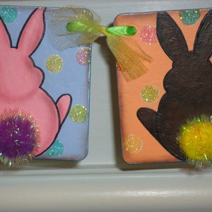 May include: Two painted wooden bunny shapes with pom-poms for tails. The bunny on the left is pink with a purple pom-pom and the bunny on the right is brown with a yellow pom-pom. Both bunnies are on a background of colorful polka dots.