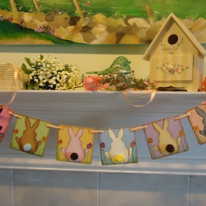 May include: A colorful Easter bunny banner with six flags featuring different colored bunnies with white tails. The banner is hanging on a white mantelpiece.