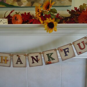 May include: A Thanksgiving banner with the word "THANKFUL" spelled out in colorful letters with fall leaves and pumpkins. The banner is hanging on a white fireplace mantel.