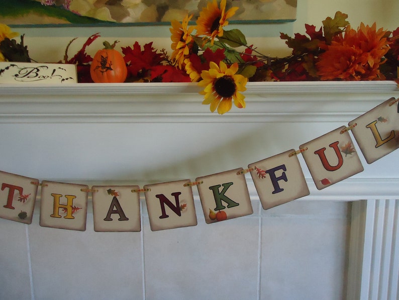 May include: A Thanksgiving banner with the word "THANKFUL" spelled out in colorful letters with fall leaves and pumpkins. The banner is hanging on a white fireplace mantel.