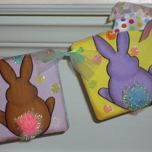 May include: A handmade Easter bunny banner with two purple and brown bunnies with pink and blue pom-poms for tails. The banner is decorated with glitter and tulle.