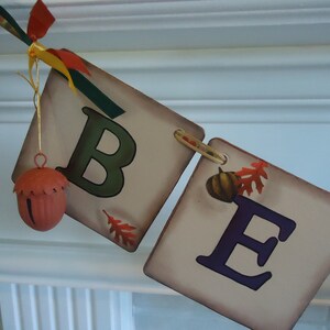May include: A fall-themed banner with the letters "BE" in green and purple paint. The banner is made of fabric and has a rustic look. There is a small acorn-shaped bell hanging from the top of the banner.