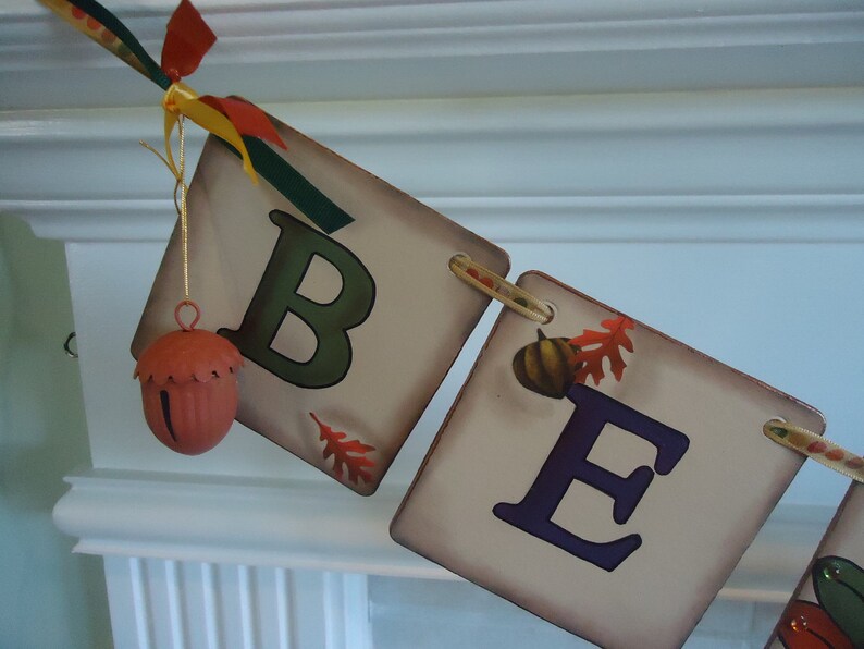 May include: A fall-themed banner with the letters "BE" in green and purple paint. The banner is made of fabric and has a rustic look. There is a small acorn-shaped bell hanging from the top of the banner.
