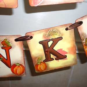 May include: A Thanksgiving banner with the letters "N" and "K" in brown, with fall-themed illustrations of leaves and pumpkins. The banner is strung together with brown ribbon.