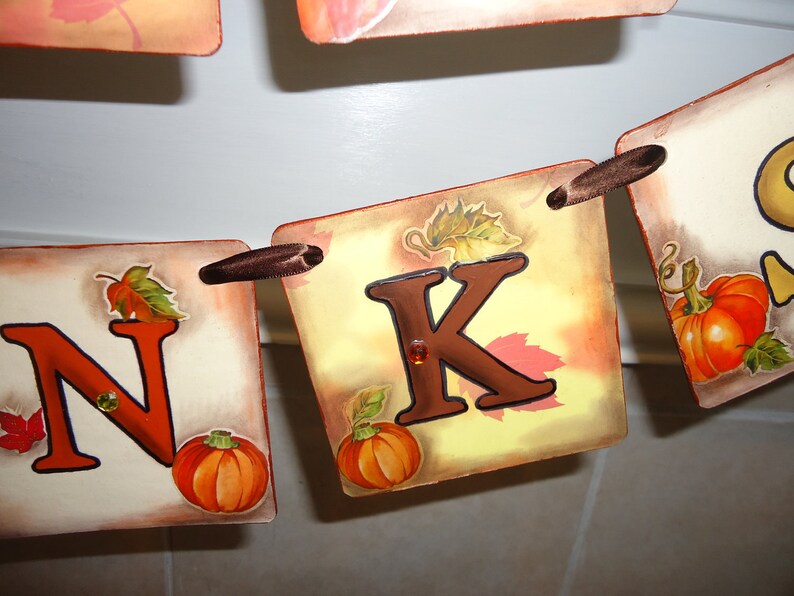May include: A Thanksgiving banner with the letters "N" and "K" in brown, with fall-themed illustrations of leaves and pumpkins. The banner is strung together with brown ribbon.