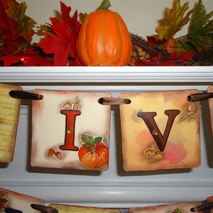 May include: A Thanksgiving banner with the word "GIVE" in brown letters on a beige background with fall leaves and acorns. The banner is strung with brown ribbon and has a pumpkin on each letter.
