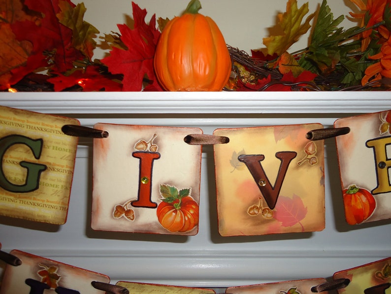 May include: A Thanksgiving banner with the word "GIVE" in brown letters on a beige background with fall leaves and acorns. The banner is strung with brown ribbon and has a pumpkin on each letter.