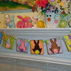 May include: A colorful Easter bunny banner with six individual flags. Each flag features a different colored bunny with a fluffy tail. The banner is strung with green and white ribbon and is displayed on a white mantel.