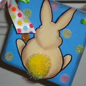 May include: A blue and white Easter bunny banner with a yellow pom-pom tail. The bunny is surrounded by white and silver glitter dots.