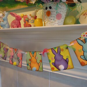 May include: A colorful Easter bunny banner with a string of flags featuring different colored bunnies with white pom-poms for tails. The banner is hanging on a white mantel.
