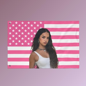 Può includere: Una bandiera americana rosa e bianca con una donna con una canotta bianca. La bandiera ha strisce rosa e stelle bianche. La donna ha lunghi capelli scuri e indossa una collana d'argento.