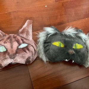 May include: Two cat-shaped masks. One is brown with darker brown stripes and blue eyes. The other is gray with yellow eyes, a brown nose, and white fur trim. Both masks have a cutout for the mouth.