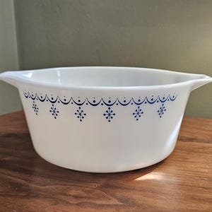 Pode incluir: Tigela de cerâmica branca com um padrão floral azul na borda. A tigela tem duas alças e está sobre uma superfície de madeira. A tigela é provavelmente vintage e adequada para servir ou expor.