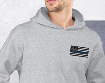 May include: A light gray hooded sweatshirt featuring a small embroidered American flag with a blue stripe. The flag design is on the upper left chest area of the sweatshirt. The hoodie has a drawstring and a front pocket.
