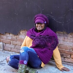 Puede incluir: Una mujer con una bufanda y un gorro de ganchillo morado, pantalones vaqueros azules y botas verdes. Est&aacute; sentada en una superficie de ladrillo con una pared negra detr&aacute;s de ella.