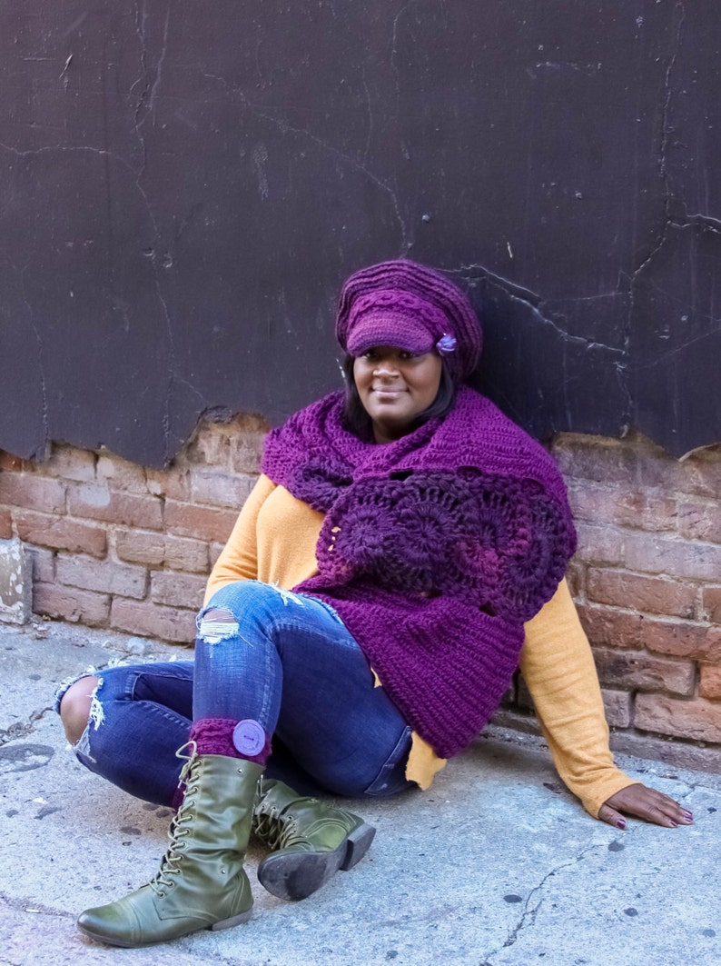Puede incluir: Una mujer con una bufanda y un gorro de ganchillo morado, pantalones vaqueros azules y botas verdes. Est&aacute; sentada en una superficie de ladrillo con una pared negra detr&aacute;s de ella.