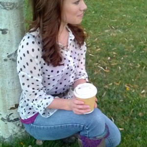 May include: A woman wearing a white and black polka dot shirt with a brown belt, blue jeans, and purple socks. She is holding a brown paper cup with a white lid. She is sitting on the ground next to a birch tree.