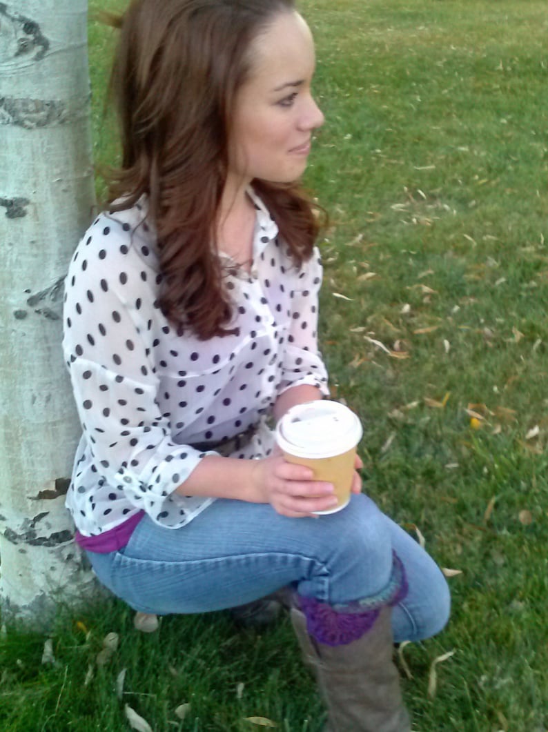 May include: A woman wearing a white and black polka dot shirt with a brown belt, blue jeans, and purple socks. She is holding a brown paper cup with a white lid. She is sitting on the ground next to a birch tree.