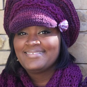 Puede incluir: Un gorro morado tejido con una peque&ntilde;a ala y una bufanda a juego. El gorro tiene un adorno decorativo en el lateral. La mujer de la imagen lleva el gorro y la bufanda.
