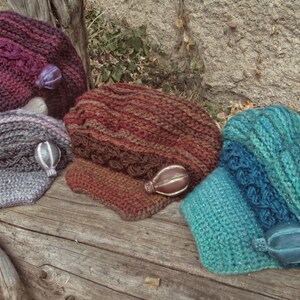 Puede incluir: Una colecci&oacute;n de sombreros tejidos a mano en varios colores, incluyendo morado oscuro, gris y turquesa. Cada sombrero presenta texturas &uacute;nicas y detalles decorativos, exhibidos sobre una superficie de madera r&uacute;stica.