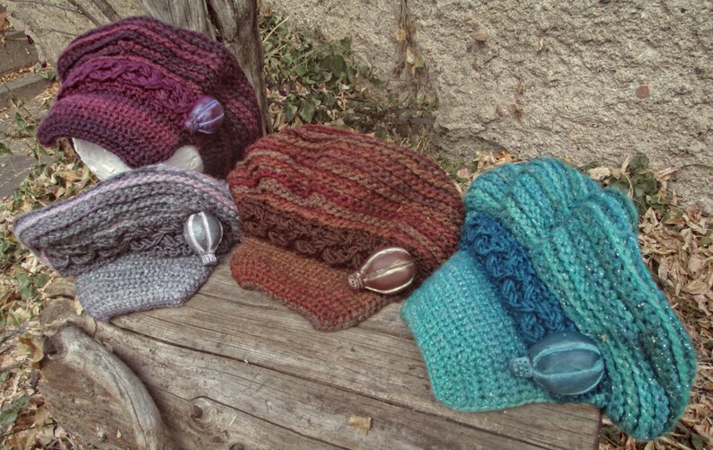 Puede incluir: Una colecci&oacute;n de sombreros tejidos a mano en varios colores, incluyendo morado oscuro, gris y turquesa. Cada sombrero presenta texturas &uacute;nicas y detalles decorativos, exhibidos sobre una superficie de madera r&uacute;stica.