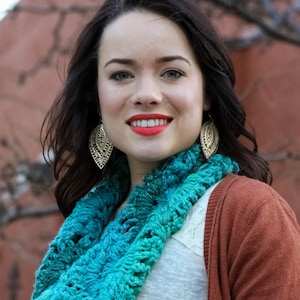 Op de afbeelding: Een vrouw met lang donker haar draagt een tealblauwe gecrochete oneindige sjaal, een wit shirt en een roestkleurige cardigan. Ze draagt gouden bladeren oorbellen.