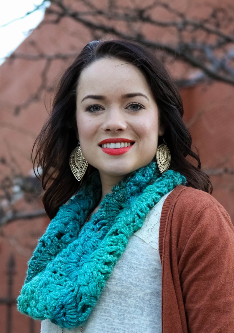 Op de afbeelding: Een vrouw met lang donker haar draagt een tealblauwe gecrochete oneindige sjaal, een wit shirt en een roestkleurige cardigan. Ze draagt gouden bladeren oorbellen.