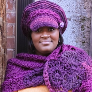 Op de afbeelding: Een vrouw draagt een paarse gehaakte hoed met een rand en een bijpassende paarse gehaakte sjaal. De hoed heeft een klein paars bloemetje als versiering.
