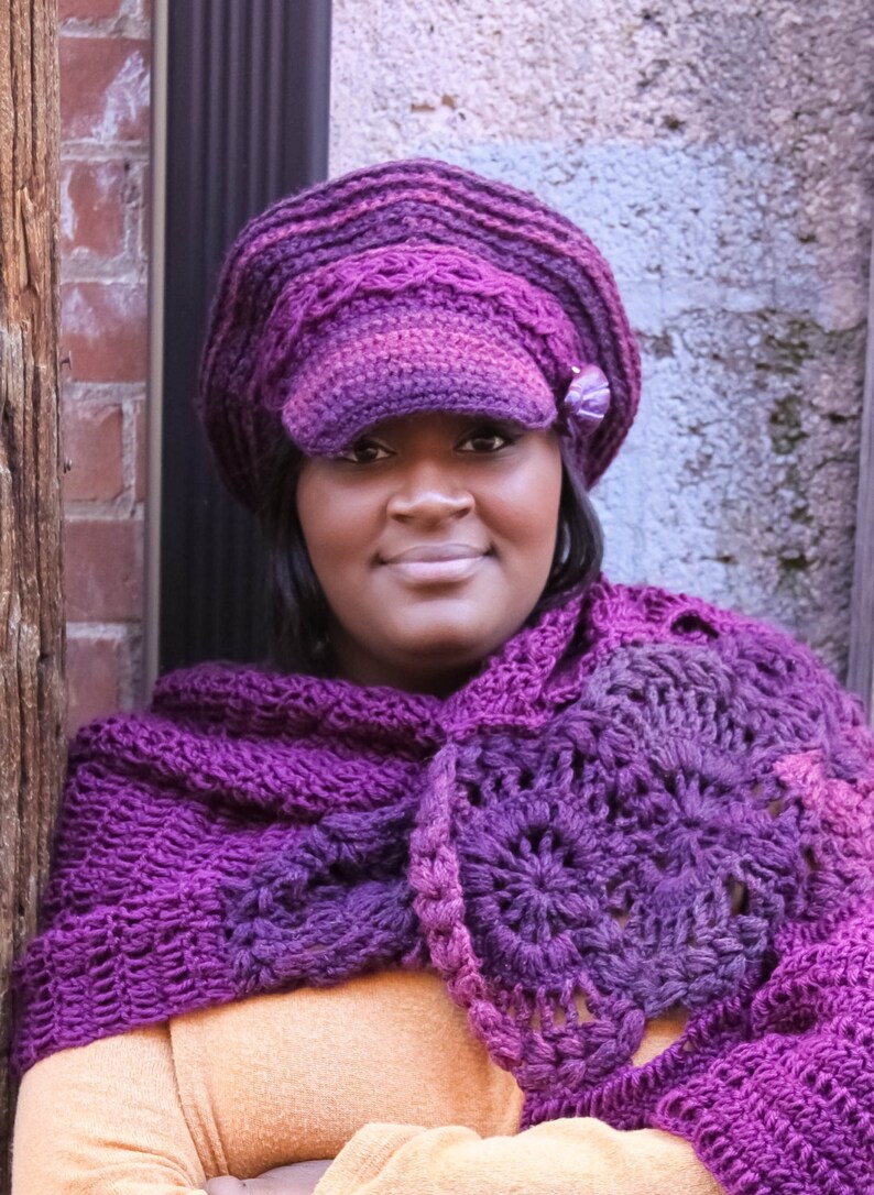 Op de afbeelding: Een vrouw draagt een paarse gehaakte hoed met een rand en een bijpassende paarse gehaakte sjaal. De hoed heeft een klein paars bloemetje als versiering.