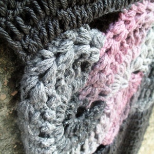 Op de afbeelding: Close-up van een gehaakte sjaal met een kleurverloop van zwart, grijs en roze. De sjaal is gemaakt van een dik garen en heeft een textuur met hobbels.