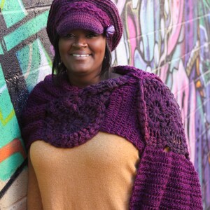 Op de afbeelding: Een vrouw draagt ​​een paarse gehaakte sjaal en een paarse newsboy-pet met een bloemenversiering. Ze staat voor een kleurrijke graffitimuur.