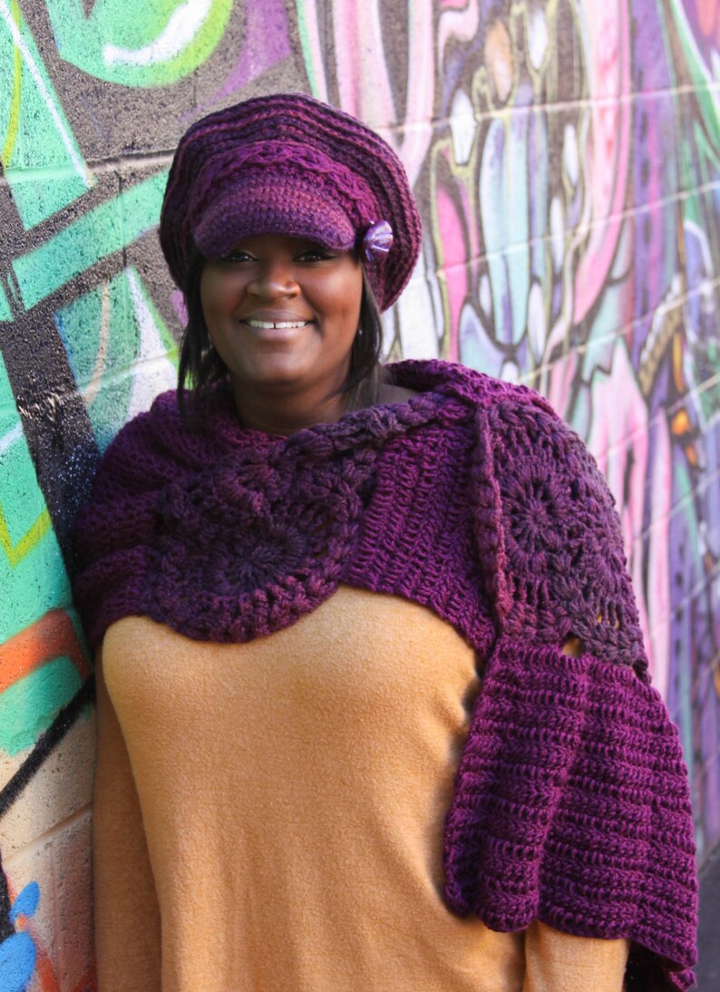 Op de afbeelding: Een vrouw draagt ​​een paarse gehaakte sjaal en een paarse newsboy-pet met een bloemenversiering. Ze staat voor een kleurrijke graffitimuur.