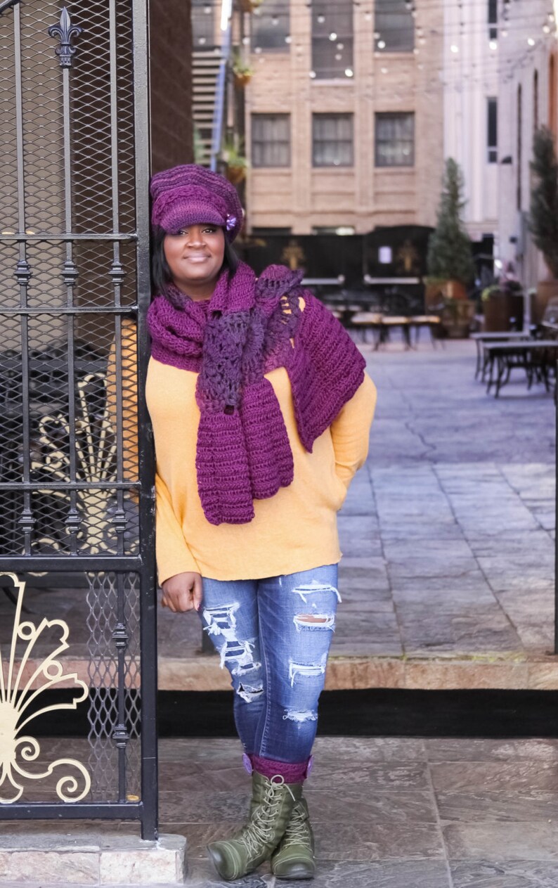 Op de afbeelding: Een vrouw draagt een paarse gehaakte sjaal en hoed, een gele trui, blauwe jeans met distressed details en groene combatlaarzen.