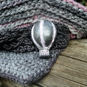 Puede incluir: Un cuello tejido a mano en gris, negro y rosa con un broche de globo aerost&aacute;tico plateado. El cuello est&aacute; hecho de hilo suave y tiene un dise&ntilde;o texturizado. El broche del globo aerost&aacute;tico est&aacute; detallado con acentos rosas.