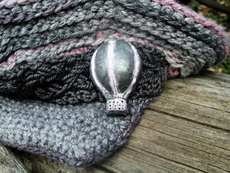 Puede incluir: Un cuello tejido a mano en gris, negro y rosa con un broche de globo aerost&aacute;tico plateado. El cuello est&aacute; hecho de hilo suave y tiene un dise&ntilde;o texturizado. El broche del globo aerost&aacute;tico est&aacute; detallado con acentos rosas.