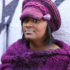 Puede incluir: Una mujer con un sombrero morado tejido a mano, con una peque&ntilde;a visera y un chal tejido a juego. El sombrero presenta un lazo decorativo en el lateral. El chal est&aacute; drapeado sobre los hombros, mostrando un dise&ntilde;o texturizado y calado.