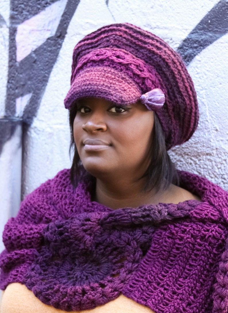 Puede incluir: Una mujer con un sombrero morado tejido a mano, con una peque&ntilde;a visera y un chal tejido a juego. El sombrero presenta un lazo decorativo en el lateral. El chal est&aacute; drapeado sobre los hombros, mostrando un dise&ntilde;o texturizado y calado.