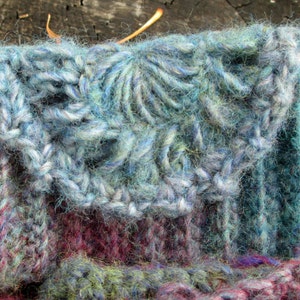 May include: Close-up of a knitted fabric with a blue and green color scheme. The fabric has a textured, fuzzy appearance and is made with a variety of yarn types.