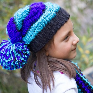 以下が含まれることがあります： 青い、紫色の、そして黒いニット帽にふわふわのポンポンをつけた女の子。帽子はかぎ針編みで、それに合うマフラーをしています。