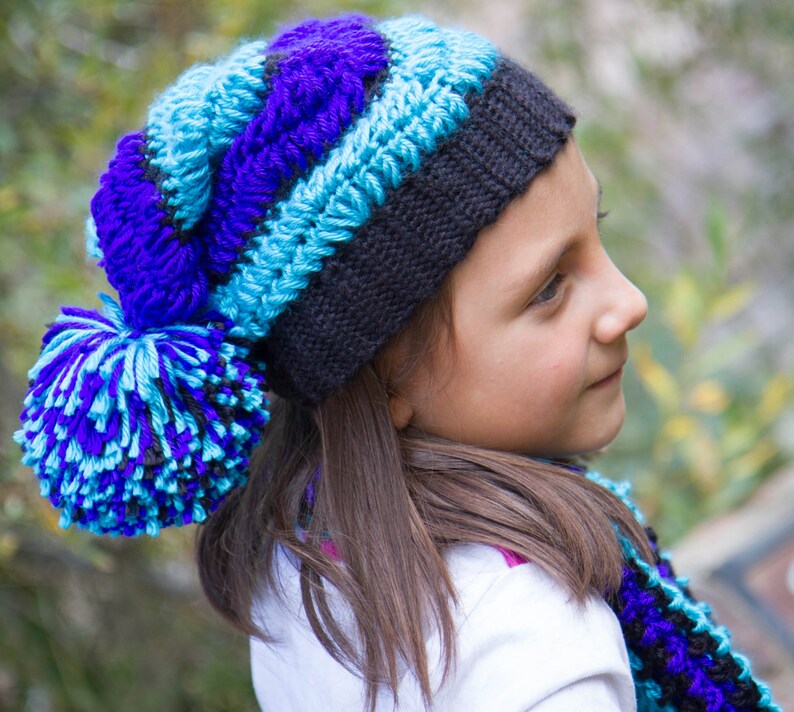 以下が含まれることがあります： 青い、紫色の、そして黒いニット帽にふわふわのポンポンをつけた女の子。帽子はかぎ針編みで、それに合うマフラーをしています。