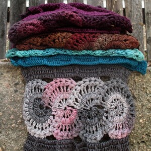 Op de afbeelding: Een gehaakt deken met een gelaagd ontwerp. De bovenste laag is donkerpaars, gevolgd door een bruine laag, een turquoise laag en een grijze laag. De onderste laag is een grijs en roze gehaakt ontwerp met een cirkelvormig patroon.