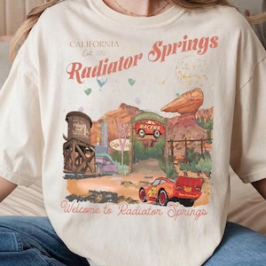 Puede incluir: Camiseta color crema con un dise&ntilde;o de Radiator Springs, con texto que dice "California Est. 2012" y "Radiator Springs". El dise&ntilde;o incluye un coche de carreras rojo y un paisaje des&eacute;rtico. "Welcome to Radiator Springs" est&aacute; impreso en la parte inferior.