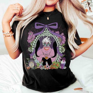Pu&ograve; includere: T-shirt nera con un'illustrazione a cartoni animati di Ursula de La Sirenetta. Il design include un fiocco viola, fiori e la scritta "Deadly Night Shade". L'opera &egrave; incorniciata all'interno di un bordo ovale.