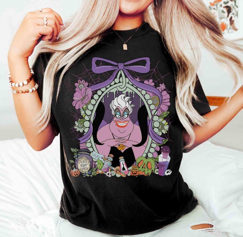 Pu&ograve; includere: T-shirt nera con un'illustrazione a cartoni animati di Ursula de La Sirenetta. Il design include un fiocco viola, fiori e la scritta "Deadly Night Shade". L'opera &egrave; incorniciata all'interno di un bordo ovale.