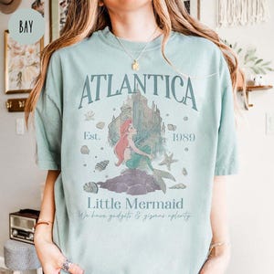 May include: A light blue t-shirt with a graphic print of Ariel, the Little Mermaid, sitting on a rock in front of a castle. The text "Atlantica Est. 1989" and "Little Mermaid We have gadgets & gizmos aplenty" is printed on the shirt.