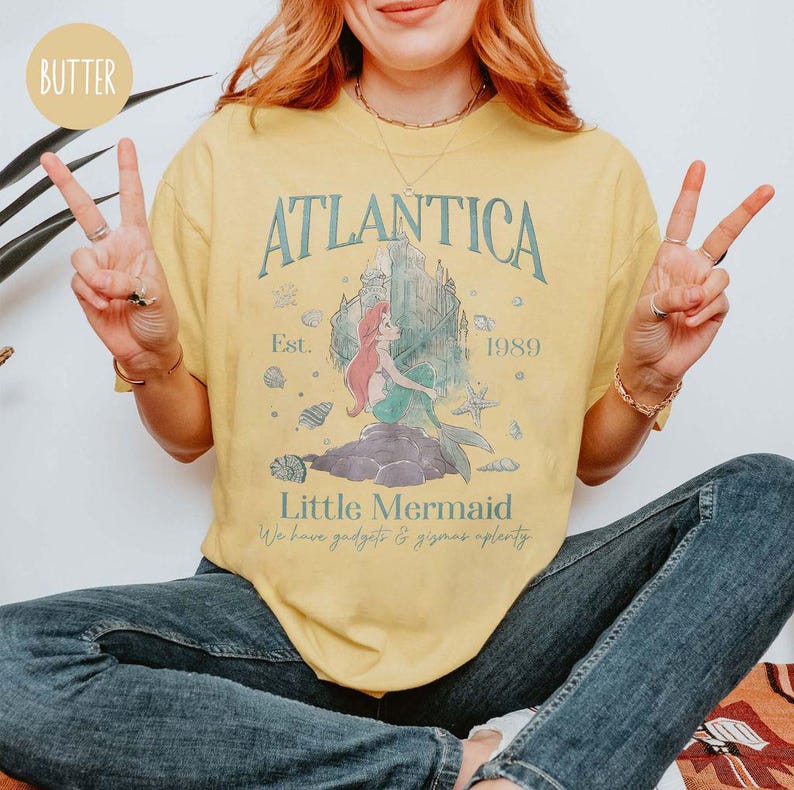 May include: A yellow t-shirt with a vintage-inspired graphic of Ariel from The Little Mermaid. The graphic features the words "Atlantica Est. 1989" and a cartoon image of Ariel sitting on a rock. The text "Little Mermaid" is printed below the graphic, along with the phrase "We have gadgets and gizmos aplenty."